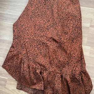 Leopardmönstrad omlottkjol Neo Noir - Snygg brun leopardmönstrad omlottkjol från Neo Noir med volangkant. Kjolen är i mjuk polyester och har knytband i midjan. Perfekt för dig som vill sticka ut med ett trendigt djurmönster och cool volangdetalj.