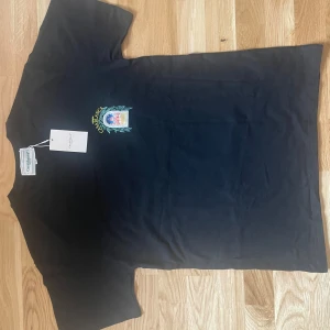 Svart t-shirt Casablanca Avenida - Helt ny Svart t-shirt från Casablanca med färgglad tryckt grafik fram och bak, där det står 'Avenida Casablanca' och ett fönster med gröna blad runt. Klassisk passform och rund hals. Materialet är mjuk bomull och t-shirten har korta ärmar. Perfekt för dig som gillar coola prints.