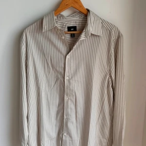 Randig loose fit skjorta från H&M - Snygg randig skjorta från H&M i loose fit-modell. Skjortan är vit med tunna gråa ränder, har klassisk krage och knäppning framtill. Långärmad och tillverkad i mjuk bomull som känns skön mot huden. Perfekt för dig som gillar en avslappnad stil.