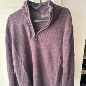 Mörklila stickad tröja Dressmann - Säljer en mörklila stickad tröja från Dressmann i storlek M. Tröjan har en ribbad hög krage med half zip-dragkedja och är långärmad. Materialet är 50% ekologisk bomull och 50% ull, vilket gör den både mjuk och varm. Perfekt för dig som gillar chill och stilren look.