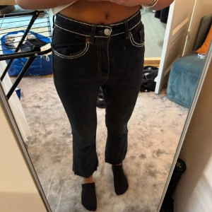 Svarta jeans med kontrastsömmar - Säljer ett par svarta jeans med vita kontrastsömmar och hög midja. Byxorna har raka ben med lätt bootcut och rå kant nedtill. Klassisk femficksmodell med knapp och dragkedja framtill. Perfekt för dig som gillar en edgy stil.