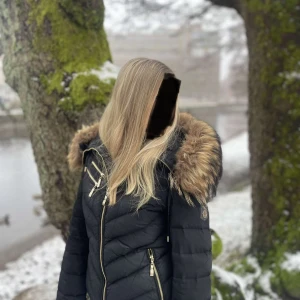 Svart pufferjacka med pälsluva - Snygg svart pufferjacka med fluffig pälsimitation runt luvan och guldfärgade dragkedjor. Jackan har quiltad design, lång ärm och flera fickor med dragkedja. Perfekt för kalla dagar och ger en cool vibe till din outfit.