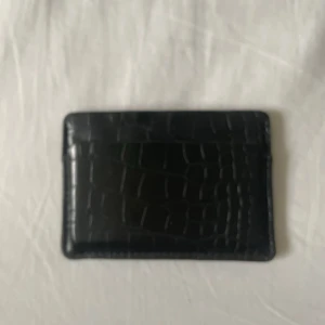 Svart plånbok i skinn med croco-mönster - Slimmad svart plånbok med croco-mönster, perfekt för kort och sedlar. Snygg och stilren design med flera fack. Materialet är skinn och ytan har en cool struktur som ger extra edge till din look.