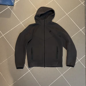 Svart Nike Tech Fleece hoodiejacka - Nike Tech Fleece hoodiejacka i svart med stilren design och hel dragkedja framtill. Jackan har två fickor med dragkedja, lång ärm och en skön huva. Materialet är mjukt och lätt, perfekt för chill eller träning. Diskret Nike-logga på bröstet för clean look.