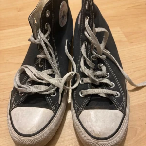 Svarta Converse Chuck Taylor High - Klassiska svarta Converse Chuck Taylor All Star high-tops med vit gummitå och sula. Skorna har vit snörning, rund tå och det ikoniska logotypmärket på sidan. Tillverkade i slitstark bomullscanvas med kontrasterande vita sömmar och detaljer.