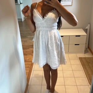 Vit spetsklänning med v-ringning - Superfin vit kortklänning med spetsdetaljer och broderat blommönster. Klänningen har v-ringad omlottfront, smala axelband och en lätt rynkad midja som ger en söt siluett. 