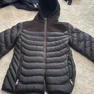Svart Moncler pufferjacka med huva - Säljer en svart pufferjacka från Moncler med broderad logga på ryggen och fluffigt foder i huvan. Jackan har snygga quiltade paneler, två dragkedjefickor och hel dragkedja framtill. Perfekt för dig som vill ha en stilren och varm jacka med exklusiv känsla. Passar både S och M