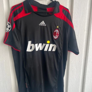 AC Milan Ronaldinho 80 Adidas fotbollströja - Snygg svart AC Milan fotbollströja från Adidas med röda detaljer och Champions League-märke på ärmen. Framsidan har Bwin-logga och klubbmärke, baksidan har Ronaldinho och nummer 80 i vitt. Klassisk krage och korta ärmar, tillverkad i lätt polyester.