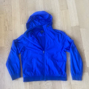Blå vindjacka från Polo Ralph Lauren - Snygg blå vindjacka från Polo Ralph Lauren i storlek XL barn, skulle säga att den passar allt mellan 165-173. Jackan har huva, hel dragkedja framtill och ribbade muddar vid ärmslut och nederkant. Perfekt för dig som gillar sportig och clean stil. Materialet är lätt och känns skönt mot kroppen.