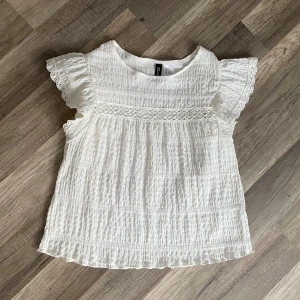 Vit blus med spets - Supersöt vit/ beige blus från H&M Divided i storlek S. Blusen har korta volangärmar med spetskant, ett smockat tyg och dekorativ spetsdetalj över bröstet. Luftig och lätt, perfekt för varma dagar. Rund halsringning och loose passform.