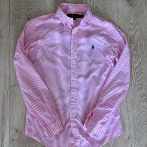 Rosa randig skjorta från Ralph Lauren - Snygg rosa och vit randig skjorta från Ralph Lauren i slim fit, strl S. Den är använd fåtal gånger och är i mycket fint skick. Pris är diskuterandet vid snabb affär! Tveka inte på att höra av dig om du har frågor!
