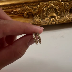 Guldiga örhängen med stenar - Säljer ett par små, runda örhängen i guldton med infattade glittrande stenar. De har en klassisk och stilren design som passar perfekt om du gillar diskreta men ändå snygga smycken. Perfekt storlek för att bära varje dag och ger en lyxig touch till din look. Köpta på en marknad i Rhodos så vet tyvärr inte materialet o därav priset!😊