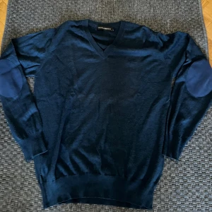 Marinblå stickad tröja Gianni Ferrucci - Snygg marinblå stickad tröja från Gianni Ferrucci med v-ringad hals och förstärkta armbågslappar i matchande blå ton. Tröjan har långärmade ribbade muddar och en klassisk passform som funkar till många olika stilar.