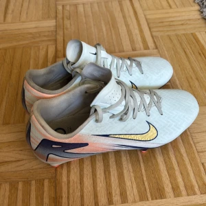 Nike Air Zoom fotbollsskor vit/guld - Säljer ett par knappt använda Nike Air Zoom fotbollsskor i vit med guldigt swoosh och detaljer i marinblått och ljusrosa. Skorna har snörning, platt sula med dobbar i korallrosa och är tillverkade i syntetmaterial. Perfekt för dig som vill ha grymt grepp och snygg design på planen. Storlek 37,5