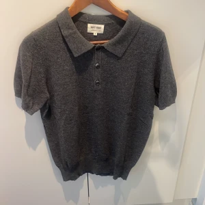 Mörkgrå kashmir pikétröja Soft Goat - Säljer en mörkgrå pikétröja från Soft Goat i 100% kashmir. Tröjan har klassisk krage, tre knappar framtill och korta ärmar. Mjuk och lyxig känsla, perfekt för dig som gillar stilrena och bekväma plagg. Passar dig som vill ha en clean look med premiumkänsla.