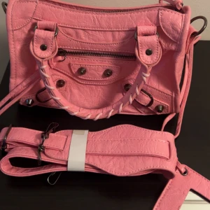 Rosa axelväska med detaljer - Säljer en snygg rosa axelväska med coola metalldetaljer och flätat handtag. Väskan har dragkedja framtill och flera nitar som ger den en edgy vibe. Medföljer en bred axelrem i samma färg och material. Perfekt för att lyfta din outfit.