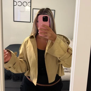 Beige croppad trenchcoat med bälten - Snygg croppad trenchcoat i beige med klassisk krage och bältdetaljer vid ärmsluten. Jackan har axelklaffar och en loose fit som ger en trendig siluett. 