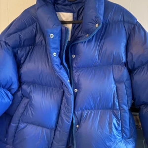 Blå pufferjacka från White:Space - Säljer en fet blå pufferjacka från White:Space i storlek L. Jackan har en hög krage, dragkedja och tryckknappar framtill samt en stor patch med loggan på ärmen. Perfekt för kalla dagar med sin fluffiga och quiltade design.