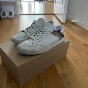 Burberry vita sneakers med rutig detalj - Snygga vita sneakers från Burberry med klassisk rutig panel i beige, svart och grått på hälen. Skorna har rund tå, platt sula och snörning framtill. Materialet är skinn och textil, och Burberry-loggan syns på plösen. Kommer med originalkartong,dustbag och kort.