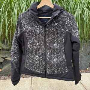 Svart mönstrad vindjacka Mountain Horse - Snygg svart vindjacka från Mountain Horse i storlek S med coolt abstrakt mönster i vitt och grått. Jackan har huva med logga, dragkedja framtill och quiltade detaljer. Perfekt för dig som gillar att sticka ut med stilren design och funktion.