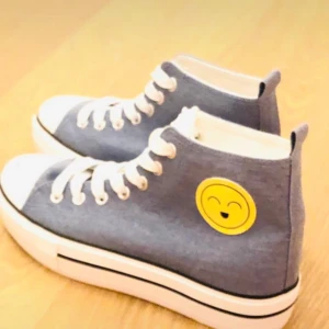 Grå höga sneakers med smiley - Säljer ett par grå höga sneakers med vit tå och vit plattformssula. Skorna har snörning och en glad gul smiley på sidan som ger en lekfull vibe. Materialet är bomullstyg och sulan är i syntet. Perfekt för dig som gillar att sticka ut med detaljer! Helt ny aldrig använd! Ingen prislapp men som ni ser aldrig används!