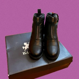 Svarta ridstövletter från B Vertigo - Snygga svarta stövletter från B Vertigo med dragkedja framtill och rund tå. Skorna är gjorda i skinn och har en platt sula med coolt mönster undertill i svart och guld. Perfekta för dig som gillar stilren design och vill ha något som sticker ut i stallet. Nya med prislapp och original förpackning. Aldrig använda original pris 2000.