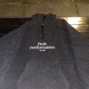 Svart fleece hoodie Peak Performance - Mjuk och varm svart hoodie från Peak Performance med broderad vit logga på bröstet. Tröjan har huva, lång ärm och half zip framtill. Tillverkad i fluffig fleece som är perfekt när det är kallt ute. Snygg och chill stil för höst och vinter.