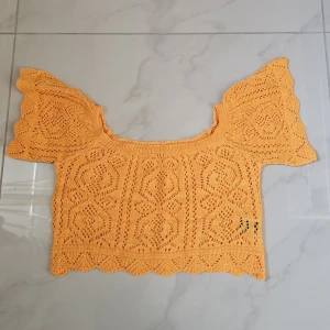 Croppad orange stickad off-shoulder topp - Superfin croppad topp i stickad orange med snyggt hålmönster och vågiga kanter. Toppen har korta, volangiga ärmar och en bred, rund halsringning. Perfekt för att ge din outfit en färgklick och cool vintagekänsla.