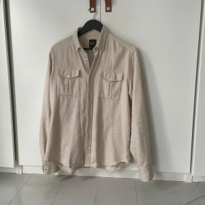 Beige overshirt från Oscar Jacobson - Snygg beige overshirt från Oscar Jacobson i storlek L med slim passform. Skjortan har två bröstfickor med knappar, klassisk krage och långa ärmar. Tillverkad i mjuk bomull som ger en chill och stilren vibe. Perfekt att bära över en t-shirt.