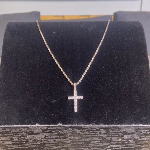 Glittrigt korshalsband i silver - Snyggt halsband med ett korsformat hänge som är täckt av glittrande stenar. Kedjan är i silverfärgad metall med tvinnad design och hänget har en riktigt clean och modern look. Perfekt för dig som vill ha något som sticker ut och glänser lite extra. Halsbandet är inte i riktigt silver eller diamant men är i ny skick.