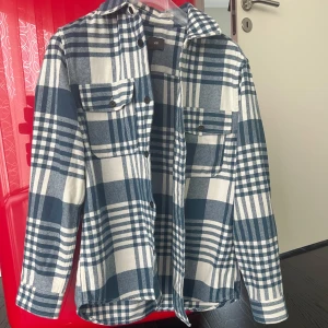 H&M - Snygg rutig overshirt från H&M i blått och vitt. Skjortan har klassisk krage, två bröstfickor med knappar och långa ärmar. Perfekt att slänga över en t-shirt för en chill look. Materialet är mjukt och känns som bomull.