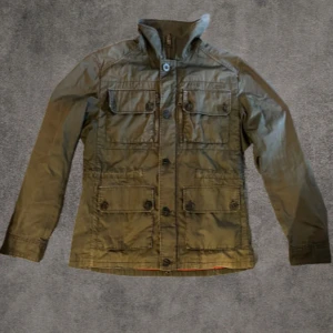 Olivgrön Fieldjacka från J.Lindeberg M - Snygg Olivgrön Fieldjacka från J.Lindeberg i storlek M. Jackan har fyra stora fickor framtill med knappar och dragkedja, klassisk krage och stilren design. Perfekt för dig som vill ha en lätt jacka med coola detaljer och funktionell look.