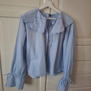 Blåvit randig blus med spetskrage - Supersöt skandinavisk blus/skjorta i ljusblått med vita ränder, spetsig krage med broderade detaljer och snörning vid halsen. Långa ärmar med knyt vid mudden ger en extra touch. Står L i storleken men funkar på S om man vill ha en lite mer oversized look