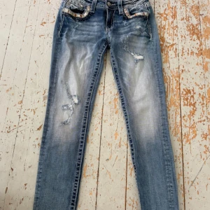 Miss Me blå skinny jeans med detaljer - Snygga blå skinny jeans från Miss Me med slitningar och broderade bakfickor med glittriga detaljer. Jeansen har låg midja, klassisk femficksdesign och coola slitningar framtill. Perfekt för dig som gillar en edgy och trendig stil.