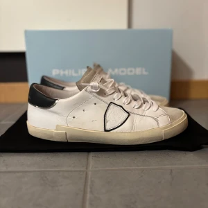 philippe models(40) - Snygga vita sneakers från Philippe Model med svart häl och beige mockadetalj på tån. Skorna har rund tå, platt sula och klassisk snörning. Ikonisk sköldlogga på sidan och stilren design som passar till jeans eller shorts. Tillverkade i skinn och mocka för en lyxig känsla.