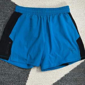 Blå träningsshorts från Silva Padel - Snygga blå träningsshorts från Silva Padel med svarta paneler på sidorna och elastisk midja. Shortsen har logga på benet och cool kontrastsöm. Perfekta för padel eller annan sport, tillverkade i lätt syntetmaterial som andas.