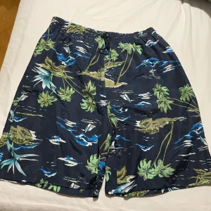 Blå tropiska badshorts från Runtech - Snygga badshorts från Runtech i storlek L med tropiskt mönster av palmer, segelbåtar och vågor i blått, grönt och vitt. Shortsen har elastisk midja med snörning och meshfoder på insidan. Perfekta för strandhäng och poolparty.