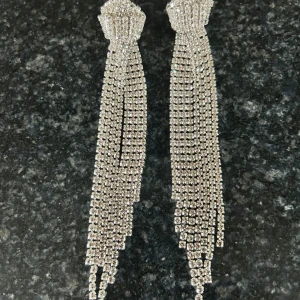 Långa glittriga silverörhängen -  örhängen i silverfärg med massor av glittrande stenar. De har flera rader av strass som hänger ner i fransar och ger en riktigt lyxig och iögonfallande look. Perfekta för att sticka ut och ge extra bling till din outfit.