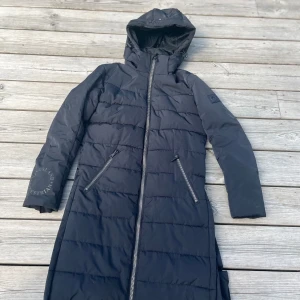 Ridkappa junior, mörkblå lång vinterjack med huva - Snygg svart lång dunjacka från Kingsland Equestrian med huva och två snedställda dragkedjefickor. Jackan har quiltad design och hel dragkedja framtill. Perfekt för kalla dagar och har stilren look med diskret logga på ärmen.