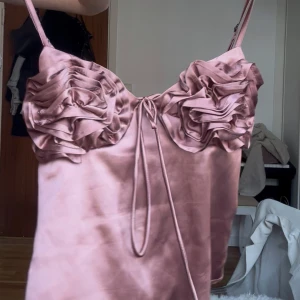 Ros topp från Zara  - Supersöt ljusrosa linnetopp i satin med justerbara smala axelband. Framsidan har stora volangdetaljer över bysten och en dekorativ knytning i mitten. Materialet är glansigt och ger en lyxig känsla. Perfekt för dig som vill sticka ut med en feminin vibe.