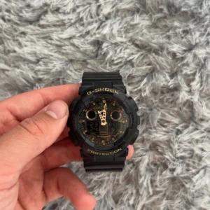 Svart G-Shock GA-100CF klocka - Säljer en fet G-Shock GA-100CF från Casio med svart resinarmband och robust boett. Urtavlan har coola detaljer i guld och digital display. Klockan är vattentät upp till 200M och har stålbaksida med G-Shock-logga. Perfekt för dig som gillar sportig och tålig stil.