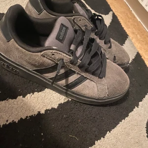 Gråa/bruna Adidas skor - Snygga gråa Adidas sneakers i mocka med svarta detaljer och klassiska tre ränder på sidan. Skorna har svart platt sula, svart snörning och rund tå. Perfekta för dig som gillar en stilren och enkel look.