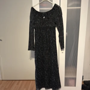 Svart långklänning med blommigt mönster - Supersnygg svart långklänning från Kappahi med diskret blommigt mönster i vitt. Klänningen har långa ärmar, rundad urringning och smock i midjan för en bekväm passform. Materialet är lätt och känns lyxigt, perfekt för dig som vill sticka ut.