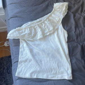 Vit oneshoulder topp med volang  - Supersöt vit oneshoulder topp från Kappahl med bred volangkant i broderad spets. Toppen är ribbad och ärmlös, perfekt för en fräsch och somrig look. Volangen har fina hålbroderier som ger en romantisk vibe. Skriv om ni har några frågor eller något o jag säljer den då den inte kommer till användning längre💕
