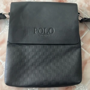 Svart axelväska från Polo Videng - Snygg svart axelväska från Polo Videng med schackrutigt mönster på framsidan. Väskan har justerbar axelrem, lock med präglad logga och dragkedja på baksidan. Tillverkad i skinnimitation och passar perfekt för att bära dina viktigaste grejer.