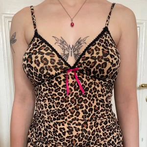 Leopardmönstrad kort klänning med rosett - Snygg kort klänning med leopardmönster och tunna axelband. Klänningen har svarta spetsdetaljer vid bysten och en rosa rosett framtill. Figurnära passform och v-ringad hals. Perfekt för dig som vill sticka ut med en djurisk look.