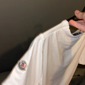 Vit slim fit t-shirt från Moncler XS - Vit t-shirt från Moncler i slim fit-modell. Klassisk design med korta ärmar, broderad Moncler-logga på ena ärmen och snygga röd-blå detaljer på ärmsluten. Tillverkad i mjuk bomull som känns skön mot huden. Perfekt för dig som gillar stilrena och exklusiva plagg. har kvitto och påse 