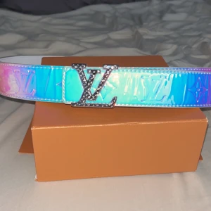 Louis Vuitton holografiskt bälte - Säljer ett Louis Vuitton bälte med holografisk yta i blå, rosa och turkosa toner. Bältet har en bred design med präglade LV-loggor och en stor, glittrig LV-spänne i silver. Kommer i originalask med blått band. Perfekt statement piece för dig som vill sticka ut. Pris diskuteras 