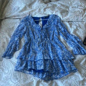 Blå blommig kortklänning från Zara - Supersöt blå och vit kortklänning från Zara med blommigt mönster, volanger och långa ärmar. Klänningen har snörning framtill, markerad midja och lätt transparent tyg med innerfoder. Perfekt för sommarens alla tillfällen!