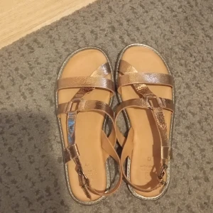 Guldiga sandaler från Next Forever Comfort - Snygga sandaler i metallic guld från Next Forever Comfort. Remmar i skinn med lätt struktur och justerbart spänne. Platt sula och öppen tå gör dem perfekta för varma dagar. Stilren design som passar till många outfits.